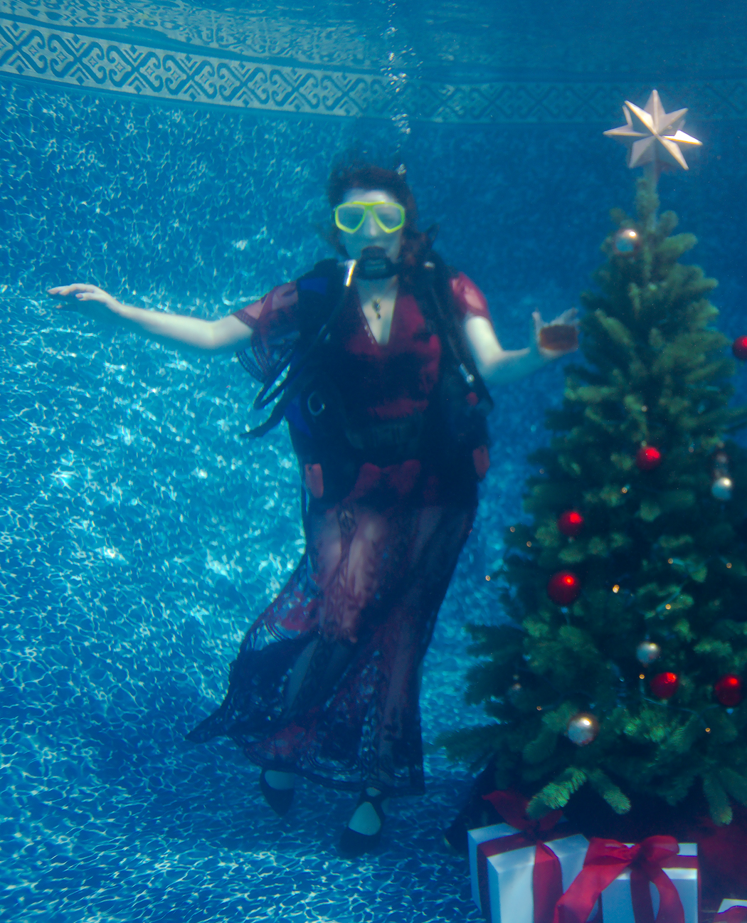A woman at the bottom of a swimming pool wearing a dress, scuba gear, and holding a glass of whiskey next to a christmas tree.